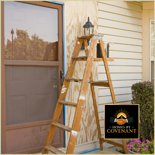 Exterior door repair in progress with ladder and plywood patch beside damaged entryway.
