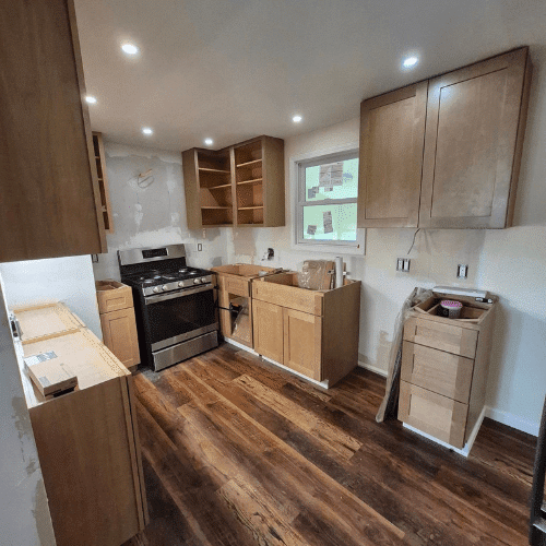 Kitchen renovation in progress showing new wood cabinetry, installed range, and partially completed backsplash under recessed lighting.